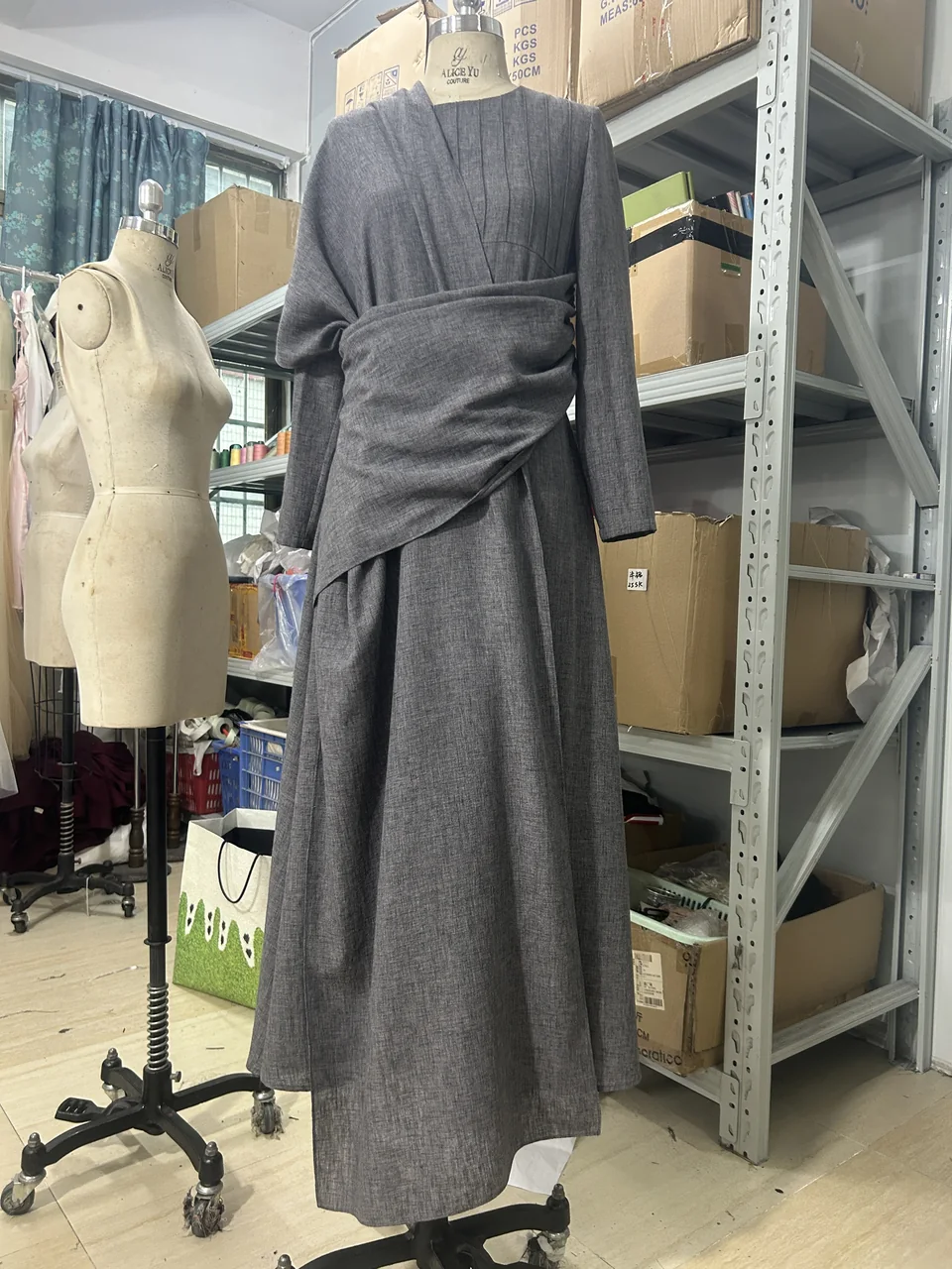 Grey long-sleeved abaya with a draped waist, photographed on a mannequin in the workspace