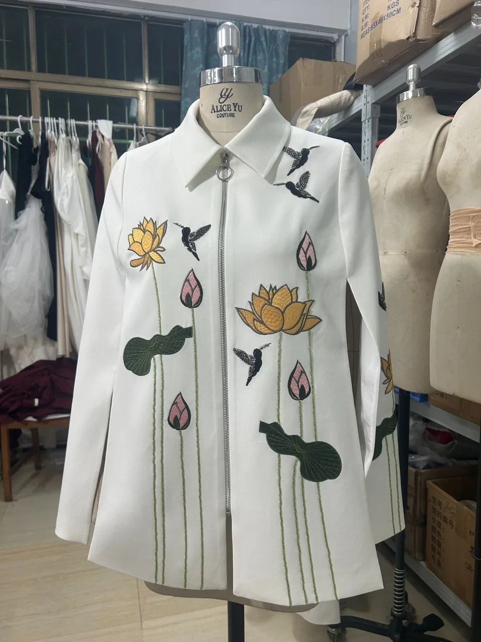 White zip-front jacket with floral embroidery, photographed on a mannequin in the workspace